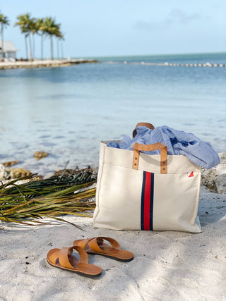 ShoreBags Natural White tote bag with navy and red stripe down the middle and natural tan handles on a beach with water in the background.