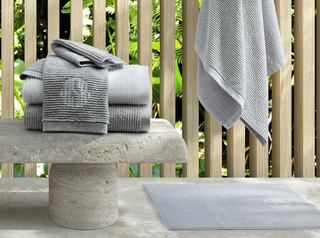 Matouk white and pool Aman towels, one with a monogram, stacked on a concrete bench with one hanging next to it.