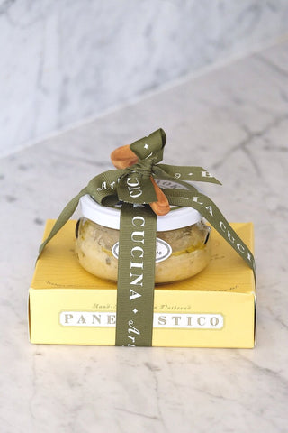 Clear glass jar of artichoke lemon pesto on top of a  yellow box of pane rustico with an olive wood spreader tied on top with an olive green Bella Cucina ribbon.